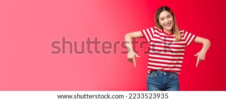 Friendly outgoing attractive blond asian girl pointing down show index fingers bottom advertisement invite guest customers try new product, smiling broadly look cheerful camera, red background.