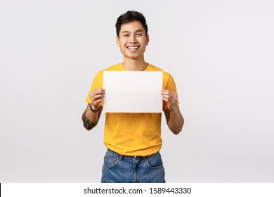 Friendly, outgoing and cheerful young asian man in yellow t-shirt, holding blank empty piece paper, smiling from overjoy, write confession or statement, showing corporate promo, white background - Powered by Shutterstock
