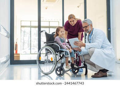 Friendly pediatrician showing someting on tablet to little patient in wheelchair. Cute preschool girl trusting her doctor and nurse in hospital. - Powered by Shutterstock