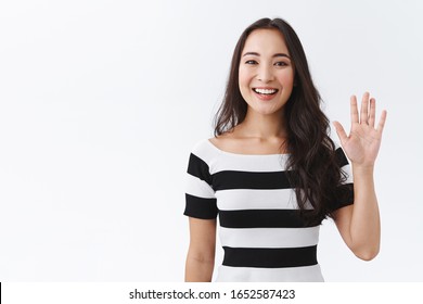 Friendly, Pleasant And Cheerful East-asian Woman In Striped T-shirt Raising Palm, Wave Hand Greeting, Saying Hi Or Hello And Smiling With Joyful Expression As Welcoming Newbies, White Background