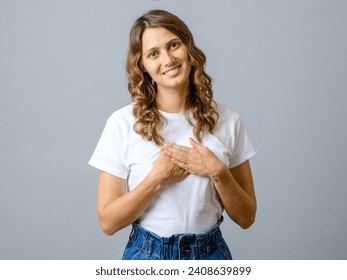 Friendly pleasant woman keeps hands on chest, touched by compliment, smiles positively over gray background