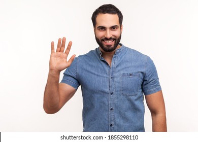 Friendly Positive Bearded Man In Blue Shirt Greeting You Rising Hand And Waving, Saying Hi, Glad To See You, Looking At Camera With Toothy Smile. Indoor Studio Shot Isolated On White Background