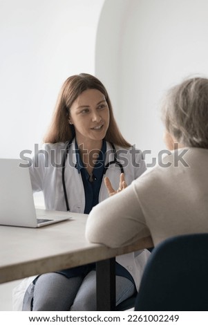 Friendly positive doctor woman seeing patient in office, speaking to elderly lady at work table with laptop, explaining checkup result, examination, treatment, making hand gestures. Vertical shot