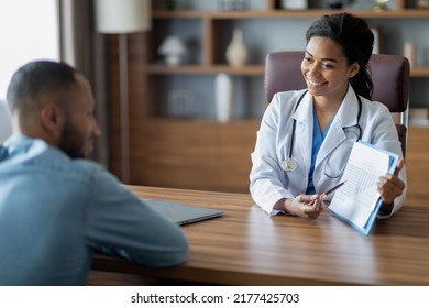 Friendly Pretty Black Lady Doctor Consulting Sick Middle Eastern Young Man Patient, Female Doc Giving Recommendations, Showing Medical Chart, Sitting At Workdesk With Laptop At Private Clinic