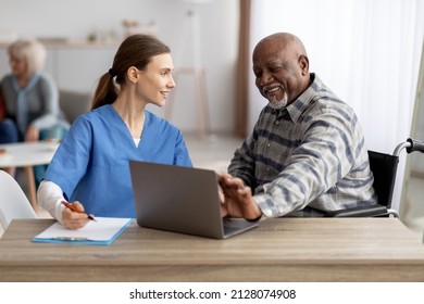 Friendly Pretty Young Woman Nurse Assisting Disabled Senior Black Man In Wheelchair Using Laptop, Sitting At Desk In Front Of Computer, Having Conversation And Smiling, Man Touching Gadget Screen