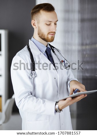 Friendly red-bearded doctor using tablet computer in clinic at his working place. Perfect medical service in hospital and medicine concept