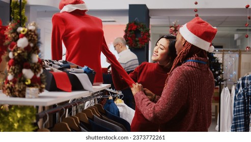 Friendly retail assistant in festive ornate shopping mall fashion shop showing client beautiful red garments, ready to be worn at Xmas themed holiday events during winter season - Powered by Shutterstock