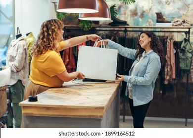 Friendly seller of clothing store giving shopping bags to satisfied female customer. Fashion store owner handing over the shopping bag to a female customer at checkout counter.