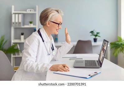 Friendly Senior Female Family Doctor Gives Online Consultation To Her Patient Via Video Call. Side View Of Doctor Sitting In Office In Front Of Laptop And Waving To Webcam. Online Doctor Concept.