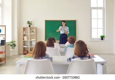 Friendly Smiley School Teacher Asks Her Elementary Students Questions In Class. Little Children Sitting At Desks And Looking At Their Teacher. Small Group Of Kids Having A Lesson In A Modern Classroom