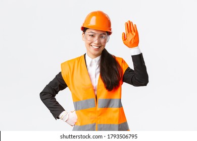 Friendly Smiling Asian Female Construction Engineer, Architect In Safety Helmet, Gloves And Glasses Waving Hand, Saying Hello, Greeting Investors Or Clients At Building Area, White Background