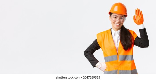 Friendly Smiling Asian Female Construction Engineer, Architect In Safety Helmet, Gloves And Glasses Waving Hand, Saying Hello, Greeting Investors Or Clients At Building Area, White Background