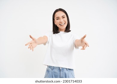 Friendly smiling asian woman, reaching her hands towards camera, hugging, welcoming you, standing over white background.