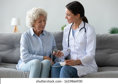 Friendly Smiling Doctor Cardiologist Physician Wearing White Uniform Checking Older Woman Blood Pressure With Digital Tonometer During Visit At Home, Sitting On Couch, Good Checkup Result