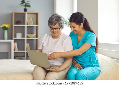 Friendly Smiling Home Care Nurse Teaching Her Senior Patient To Use Modern Computer. Happy Mature Woman Looking At Laptop Screen Watching Funny Video Or Learning To Have Video Chat With Relatives