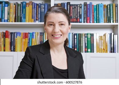 Friendly Smiling Mature Woman With Black Blazer In An Office With Lots Of Books, Professor, Teacher, Translator, Lawyer, Trustee, Accountant Or Businesswoman