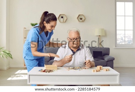 Friendly smiling nurse caregiver in geriatric clinic or retirement home helps senior man with Alzheimer's disease. Happy old man plays puzzles while sitting at table in cozy interior. Dementia concept
