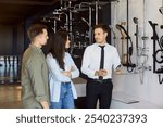 Friendly smiling salesman talking to customers, explaining differences and helping them to choose shower head for their bathroom. Young couple standing next to sales assistant at building market.