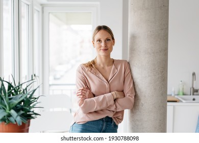 Friendly smiling young woman relaxing in an apartment kitchen leaning against an interior pillar with crossed arms looking at camera in a high key portrait with copyspace - Powered by Shutterstock