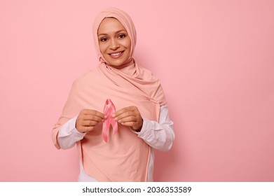 Friendly and solidar Arab Muslim woman in a pink hijab holds a pink ribbon to her chest levels for a breast cancer campaign, promoting cancer awareness. Pink October month , women's health concept - Shutterstock ID 2036353589