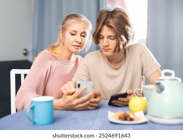 Friendly teenager helping interested mother to deal with smartphone while sitting together at table at home.. - Powered by Shutterstock