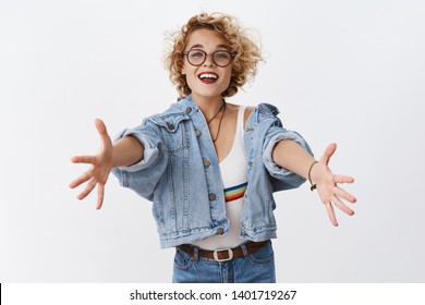 Friendly and tender outgoing cute female with short blond curly hairstyle in glasses and denim jacket stretching arms towards camera in greeting and warm welcome, wanting hug or cuddle over white wall