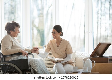 Friendly Volunteer Reading A Book To A Disabled Senior Woman In A Hospice