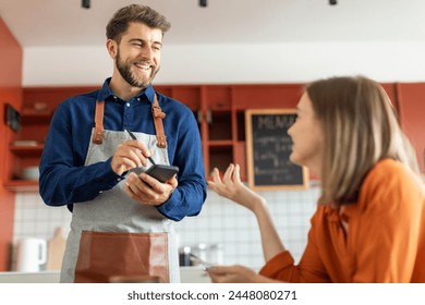 Friendly waiter taking order at the table, looking at female client and smiling, cafe interior - Powered by Shutterstock