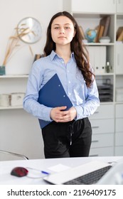 Friendly Woman With Blue Folder Of Documents Stands In Bright Office