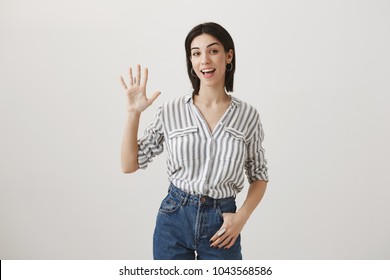 Friendly woman met her girlfriends and ready to go shopping. Studio shot of attractive girl raising one hand, waving and showing hi or hello gesture, being pleased to see familiar face in crowd