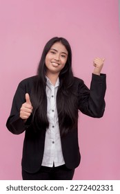 A friendly woman points to the right while giving a thumbs up gesture. Recommending a service or product. Isolated on a pink background. - Shutterstock ID 2272402331
