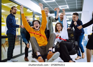 Friendly Work Team  Ride Chairs In Office Room Cheerfully Excited Diverse Employees Laugh While Enjoying Fun Work Break Activities, Creative Friendly Workers Play A Game Together.