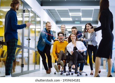 Friendly Work Team  Ride Chairs In Office Room Cheerfully Excited Diverse Employees Laugh While Enjoying Fun Work Break Activities, Creative Friendly Workers Play A Game Together.