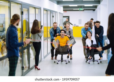 Friendly Work Team  Ride Chairs In Office Room Cheerfully Excited Diverse Employees Laugh While Enjoying Fun Work Break Activities, Creative Friendly Workers Play A Game Together.