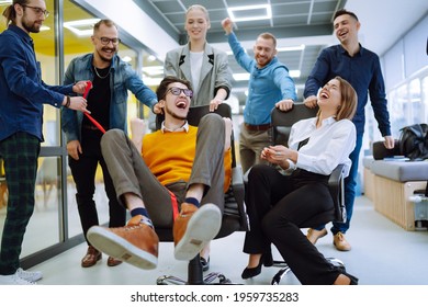 Friendly Work Team  Ride Chairs In Office Room Cheerfully Excited Diverse Employees Laugh While Enjoying Fun Work Break Activities, Creative Friendly Workers Play A Game Together.