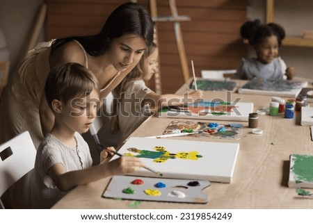 Friendly young art teacher giving artistic lesson to kids, helping to draw colorful cartoon animals, blend colors on palette, painting on canvas. Diverse group of children training creativity