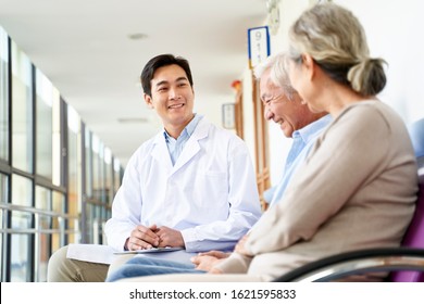 Friendly Young Asian Doctor Talking To Old Couple In Hospital Hallway