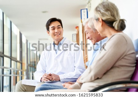 friendly young asian doctor talking to old couple in hospital hallway