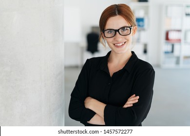 Friendly young businesswoman wearing glasses standing with folded arms giving the camera a confident smile in a spacious office