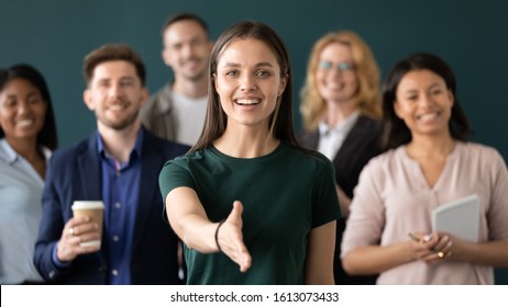 Friendly young company woman representative holds out her hand for handshake welcoming customer smiling looking at camera posing together with diverse colleagues, sales manager greeting client concept - Shutterstock ID 1613073433
