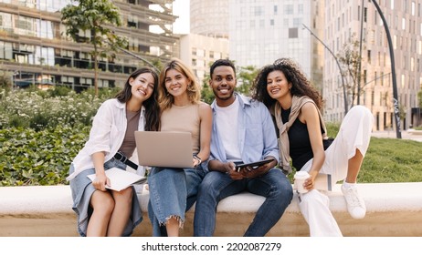 Friendly Young Diverse College Students Enjoy Modern Technology Sitting Outside. Guy With Girls Look At Camera, Wear Casual Clothes. Leisure Concept