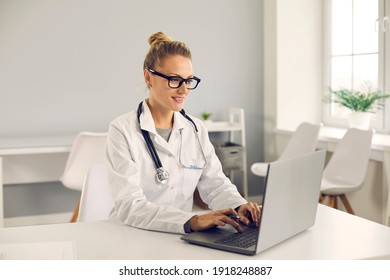 Friendly young doctor sitting at desk, using laptop computer, doing medical Internet research, entering electronic health records into database or giving online consultation to patient on eHealth chat - Powered by Shutterstock