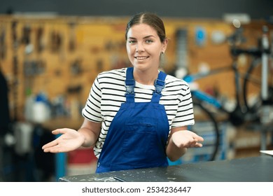 Friendly young female mechanic in blue overalls standing at service counter with open gesture, ready to provide assistance and advice in well-stocked bike repair shop - Powered by Shutterstock