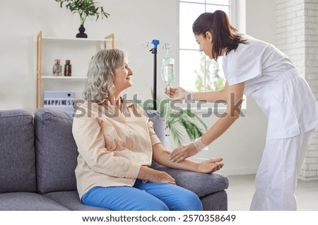 Friendly young female medical nurse helping senior elderly woman patient sitting on sofa in nursing home in receiving intravenous IV drip infusion and vitamin injection therapy to boost immune system.