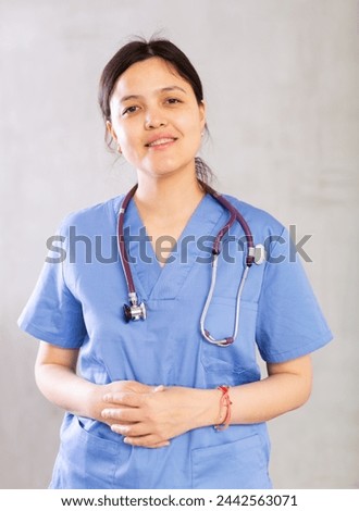 Friendly young female professional doctor in blue scrub with phonendoscope around neck posing against gray studio background, looking at camera with smile..