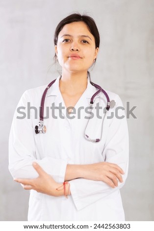 Friendly young female professional doctor in white coat with phonendoscope around neck posing against gray studio background, looking at camera with smile..