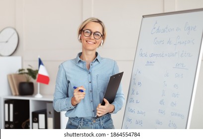 Friendly Young Female Tutor Standing Near Blackboard With French Grammar Rules, Giving Web Lesson From Home. Foreign Language Professor Teaching Students Online. Remote Education Concept