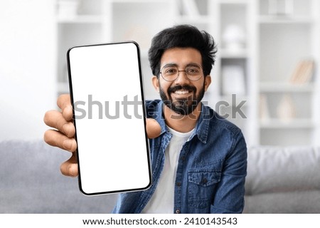 Friendly young indian man with glasses and beard smiling while presenting blank smartphone screen, handsome eastern guy sitting on couch in living room, recommending new app or website, mockup