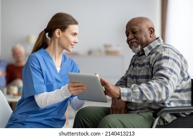 Friendly Young Lady In Workwear Nurse Helping Black Senior Man In Weheelchair Using Modern Digital Tablet, Elderly African American Male Patient Surfing On Internet At Nursing Home