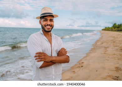 Friendly Young Latin American Man With Arms Crossed On The Beach, Wearing A Hat